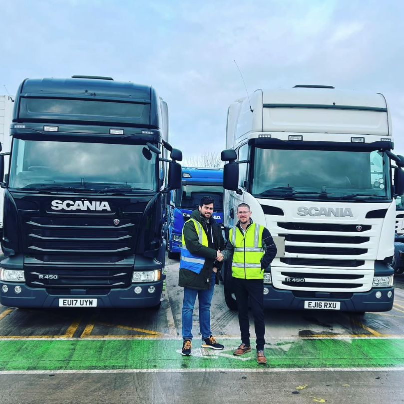 Two workers in high visibility clothing standing between a blue and a white Scania transport truck